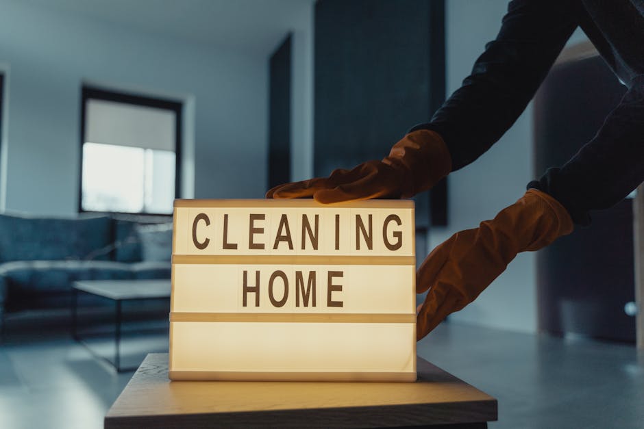 A person wearing brown gloves is adjusting a lit-up sign on a table that reads 'CLEANING HOME' in a modern, well-lit living room. The background features a dark-colored sofa, a black coffee table, and