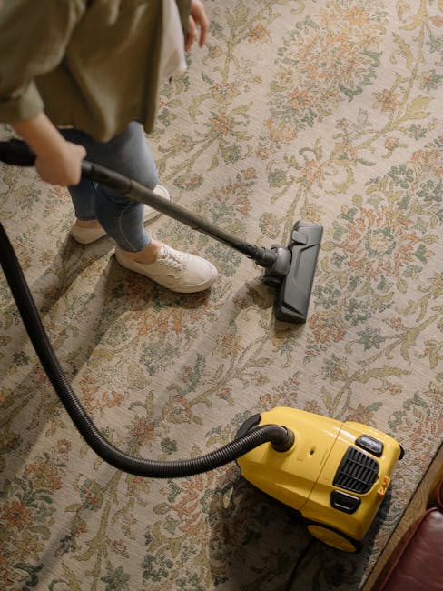 A person using a handheld vacuum cleaner to deep clean a patterned area rug in a domestic living room. The individual is dressed in casual clothing and white sneakers, with the vacuum hose connected to a yellow canister vacuum placed on the floor. The rug features a floral design with shades of green, orange, and cream, and the room is well-lit, highlighting the clean and well-maintained surface. The scene exemplifies thorough surface cleaning and sanitisation by Carpet Cleaners Barnet, in alignment with their same-day carpet cleaning services in High Barnet EN5, BARNET.