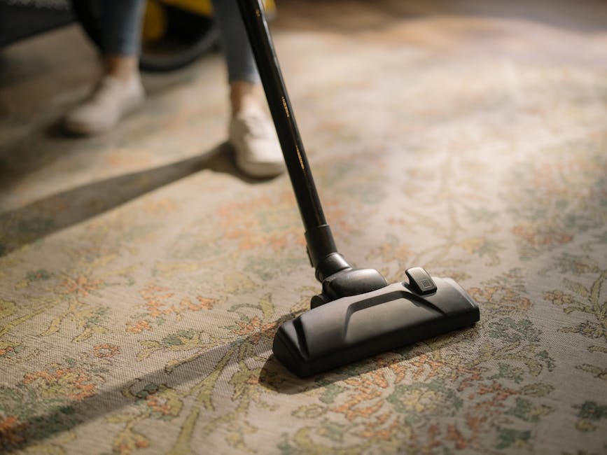 Close-up of a vacuum cleaner head actively cleaning a detailed, patterned area rug in a room with warm lighting. The area rug features an intricate floral design in muted tones of beige, green, and orange. The vacuum cleaner, which appears modern and lightweight, is positioned centrally on the rug, with a visible power button on the head. A person, wearing dark trousers and white shoes, is partially visible in the background, standing beside the vacuum. The scene emphasizes surface cleaning and deep cleaning processes aimed at maintaining hygiene and freshness in a domestic setting, consistent with the services offered by Carpet Cleaners Barnet.