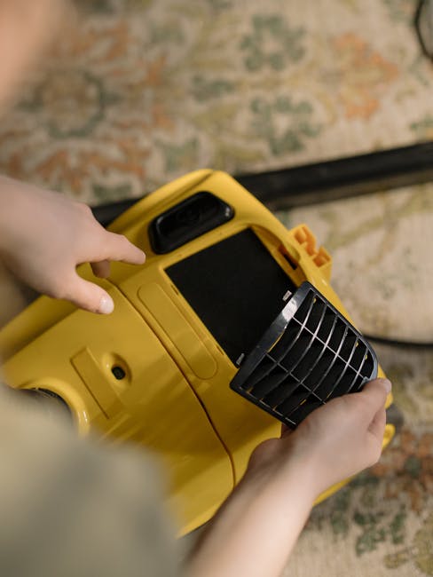 A person using a handheld vacuum cleaner to deep clean a patterned area rug in a domestic living room. The individual is dressed in casual clothing and white sneakers, with the vacuum hose connected to a yellow canister vacuum placed on the floor. The rug features a floral design with shades of green, orange, and cream, and the room is well-lit, highlighting the clean and well-maintained surface. The scene exemplifies thorough surface cleaning and sanitisation by Carpet Cleaners Barnet, in alignment with their same-day carpet cleaning services in High Barnet EN5, BARNET.